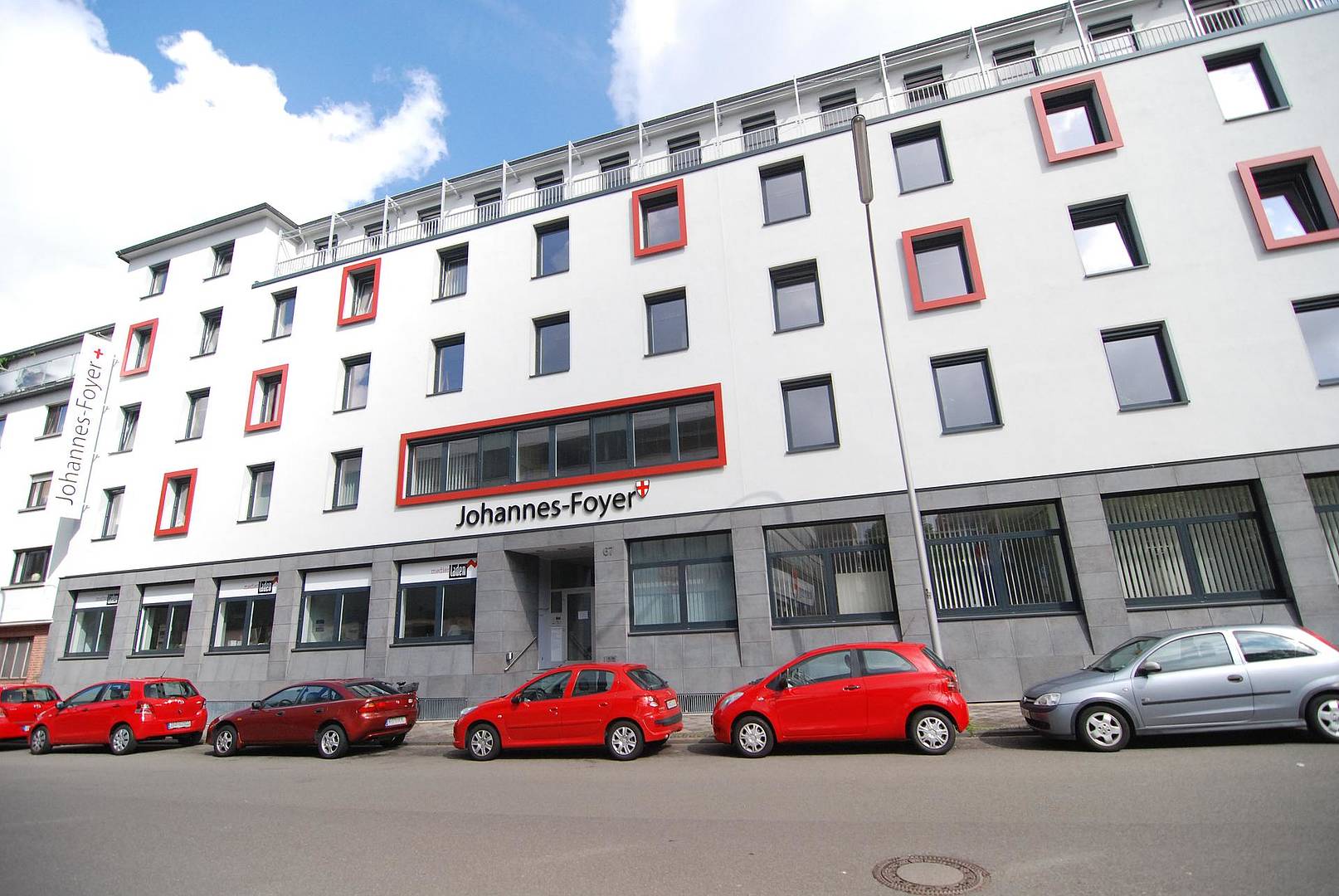 Ein großes weißes Haus mit vielen Fenstern. Einige der Fenster sind rot umrahmt. Vor dem Haus stehen einige rote Autos